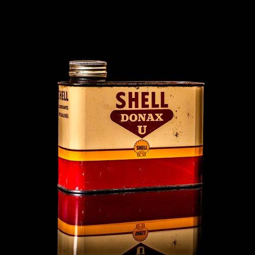Vintage Shell oil can from 1950–1960 with white base color, yellow and red accents, and the classic Shell logo. Antique motor oil tin displayed in a studio setting against a solid black background."